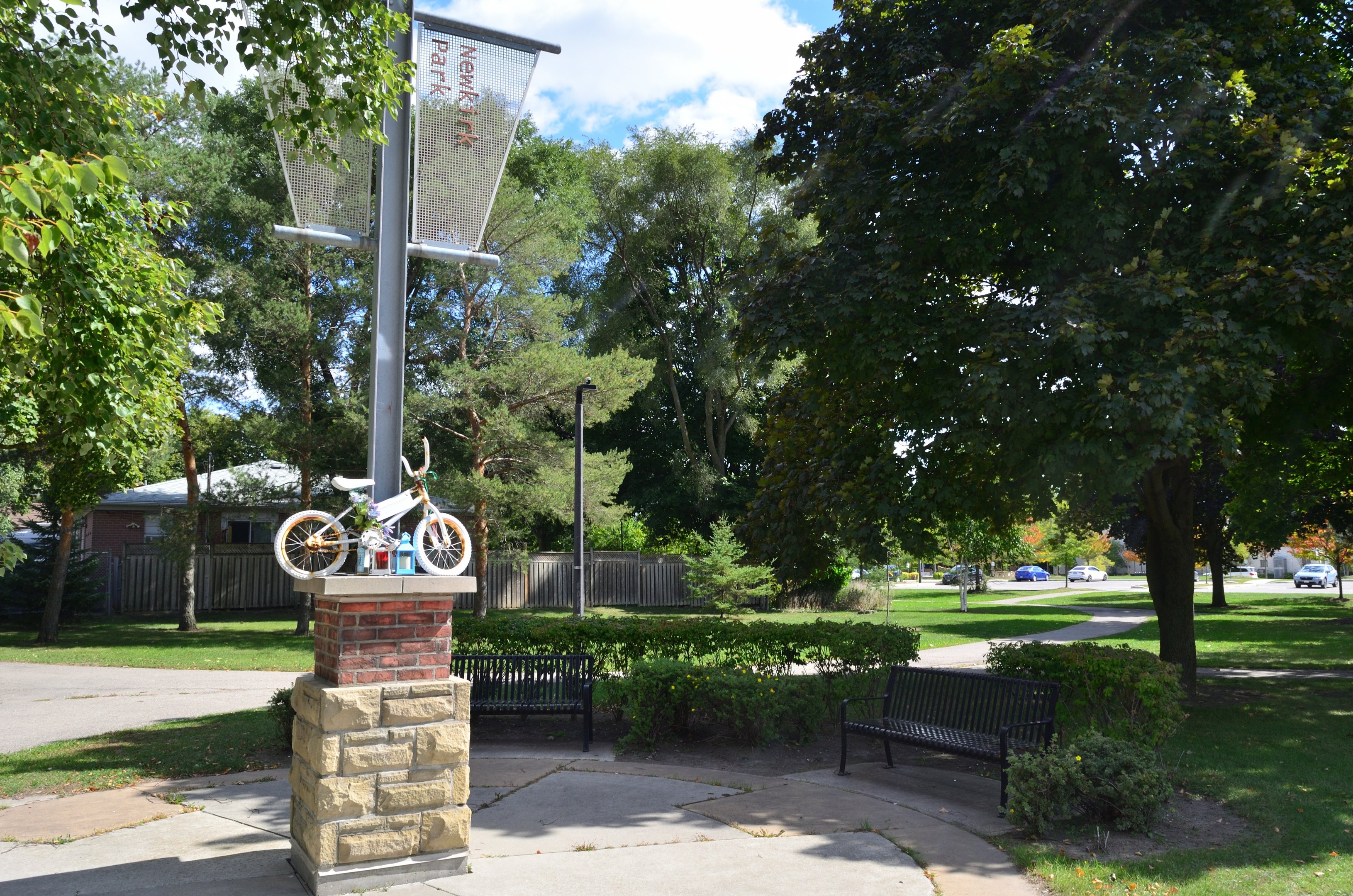 Ein Park mit einer Bank, einem Basketballkorb und einem Fahrrad auf einem zentralen Pfahl, umgeben von Bäumen, Pflanzen, Gras und Häusern, mit Fahrzeugen auf einer Straße im Hintergrund unter einem bewölkten Himmel.