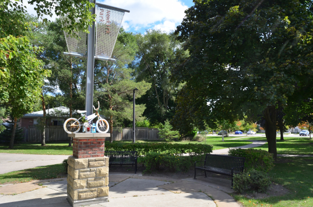 Ein Park mit einer Bank, einem Basketballkorb und einem Fahrrad auf einem zentralen Pfahl, umgeben von Bäumen, Pflanzen, Gras und Häusern, mit Fahrzeugen auf einer Straße im Hintergrund unter einem bewölkten Himmel.