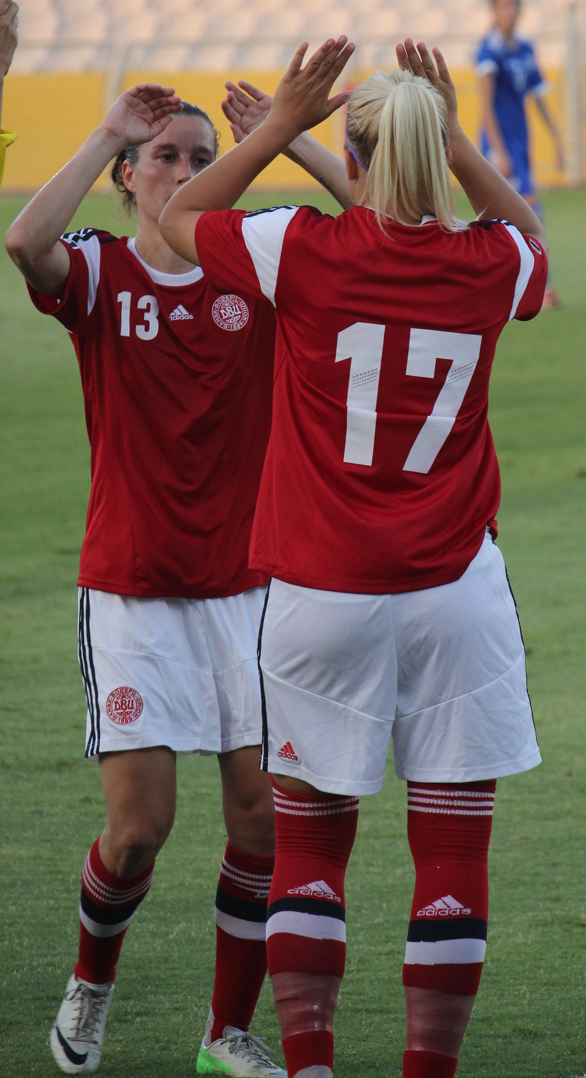 Zwei Frauen in roten T-Shirts und weißen Shorts klatschen auf einem Fußballfeld, mit einer Person in einem blauen Kleid, einer gelben Tafel und Stühlen im Hintergrund.