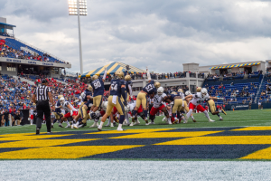 Ein Fußballspiel im Gange mit Spielern in Sportbekleidung und Helmen im Vordergrund, Zuschauern im Hintergrund und Stadionelementen wie Stühlen, Geländern, Schildern, Lichtmasten und Wolken.