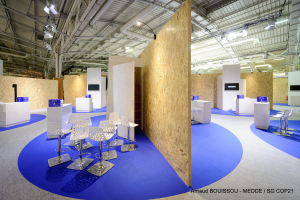 Ein Messestand mit einem blauen Teppich, weißen Stühlen, Tischen, Bildschirmen, Lampen, Stangen und anderen Gegenständen, mit einem Wasserzeichen unten, das 'Mede Sg Cop21' lautet.