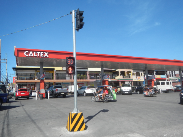 Tankstelle mit parkenden Autos und Motorrädern, Verkehrsampel im Vordergrund, Gebäude und Versorgungsmasten im Hintergrund unter wolkenlosem blauem Himmel.