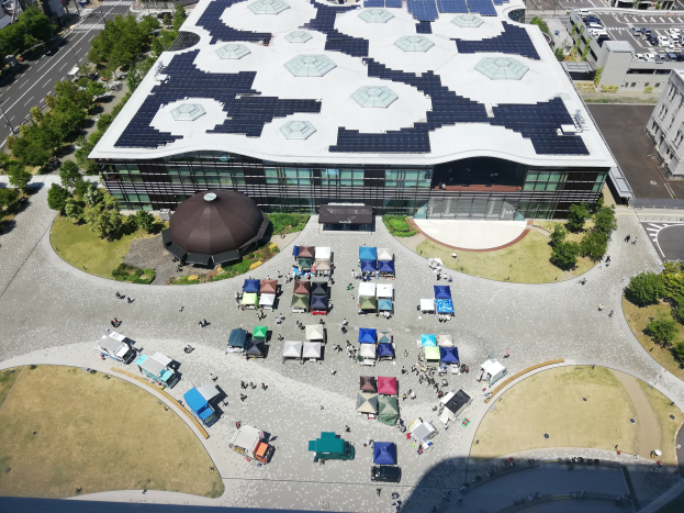 Luftaufnahme der größten Solarstromanlage Japans im Jahr 2020, die ein großes Gebäude mit Solarmodulen auf dem Dach zeigt, umgeben von Bäumen, Fahrzeugen auf der Straße und einer Gruppe von Menschen.