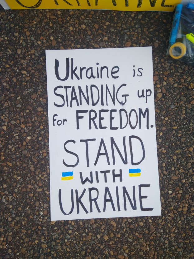 Ein weißer Zettel mit der Aufschrift "Ukraine steht für Freiheit ein, stellt dich Ukraine" vor einem gelben Banner auf dem Boden, rechts daneben eine Plastikabdeckung mit verschiedenen Gegenständen.