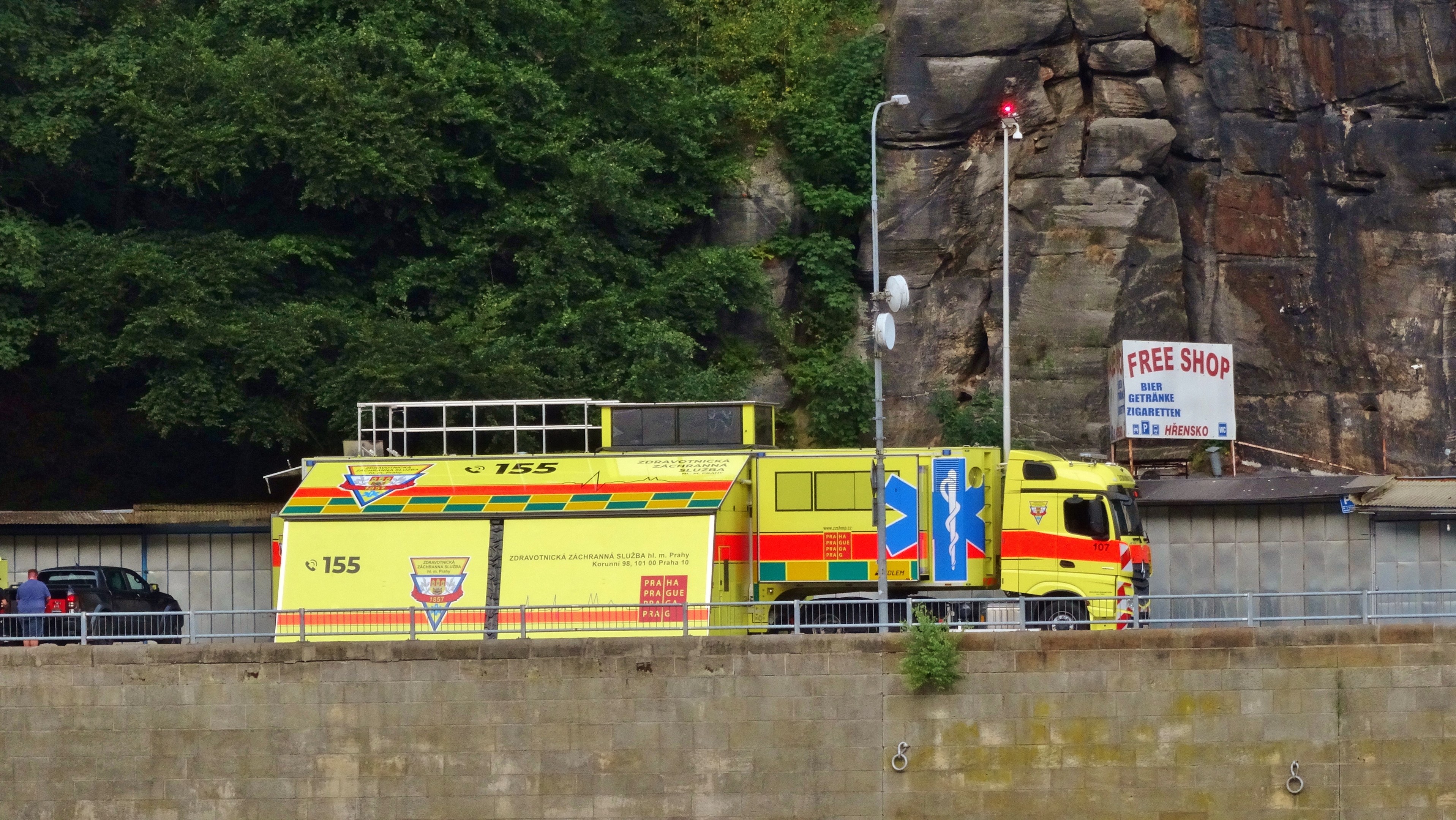 Ambulanz auf der Seite einer Straße neben einer Klippe geparkt, mit verschiedenen Gegenständen im Vordergrund und Bäumen und einem Auto im Hintergrund.