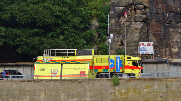 Ambulanz auf der Seite einer Straße neben einer Klippe geparkt, mit verschiedenen Gegenständen im Vordergrund und Bäumen und einem Auto im Hintergrund.