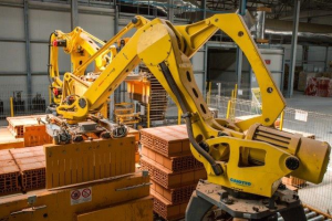 Großes industrielles Lagerhaus mit gestapelten Kisten, Kränen an der Decke, Zaun, Versorgungsmasten und einer fernen Wand in einer Fertigungseinrichtung.