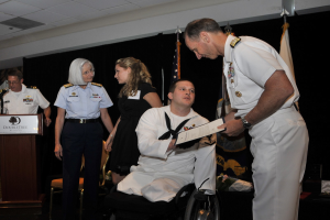 Ein Mann im Rollstuhl erhält eine Auszeichnung von einem stehenden Mann, während andere beobachten, mit einem Podium, Mikrofon und einer Tafel mit Text im Hintergrund, Flaggen und ein schwarzer Vorhang.