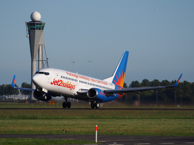 Ein Airbus A320-200 der Jet2holidays startet vom Frankfurter Flughafen, umgeben von grünem Gras und einem hohen Turm im Hintergrund, mit Bäumen und Gebäuden in der Ferne unter einem klaren blauen Himmel.