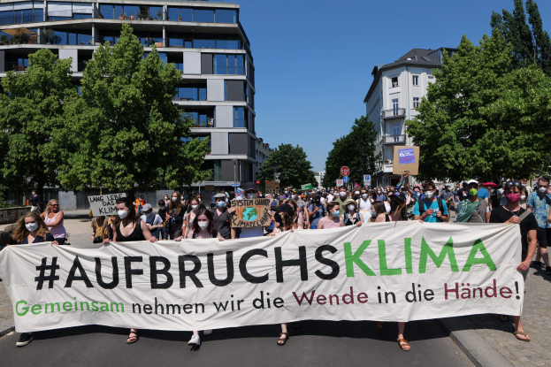 Eine Gruppe von Menschen in Masken, die ein Banner mit der Aufschrift 'Aufbruchsklima' vor einem Gebäude mit Bäumen und einem klaren Himmel halten.