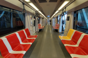 Rote und gelbe Zugsitze mit Griffen und Stangen, Glasfenster und Deckenleuchten; Passagiere sind im Hintergrund durch die Fenster sichtbar.