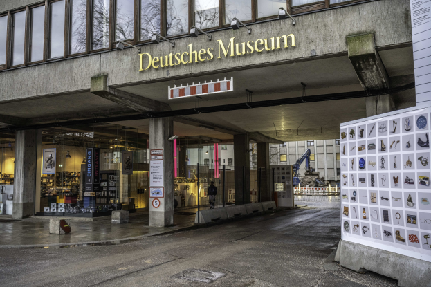 Außenansicht des Deutschen Museums in Berlin mit einem großen Gebäude mit Glasfenstern, Säulen und einem Namensschild sowie einer informativen Anzeigetafel und umliegenden städtischen Elementen.