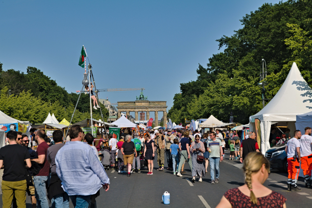 Eine Menschenmenge, die eine Straße mit Zelten, Fahrzeugen und Bäumen entlanggeht, mit einem Bogen und einem klaren blauen Himmel im Hintergrund und Polen mit Fahnen auf der linken Seite, die wahrscheinlich das Oktoberfest in München, Deutschland, darstellen.