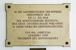 Plaque auf einem Gebäude mit der Aufschrift "In der Winterreitschule der Hofburg" auf einem hellblauen Hintergrund mit schwarzem Rand und fetter schwarzer Schrift.