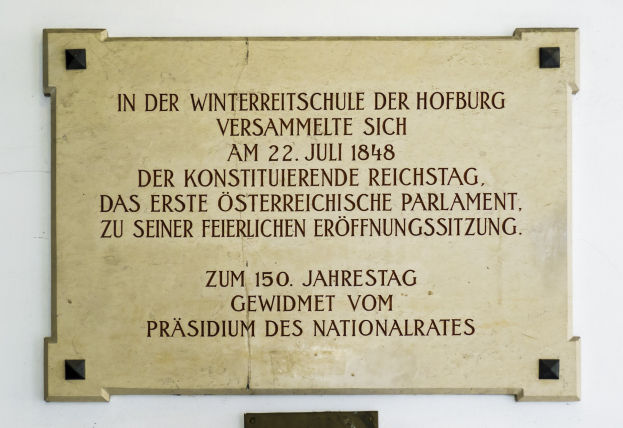 Plaque auf einem Gebäude mit der Aufschrift "In der Winterreitschule der Hofburg" auf einem hellblauen Hintergrund mit schwarzem Rand und fetter schwarzer Schrift.