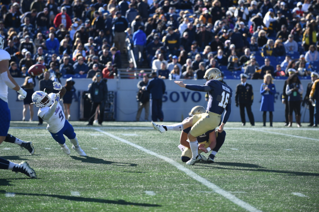 Ein Fußballspieler in der Luft einen Ball auf einem Feld mit Zuschauern in Sportkleidung und Stadion-Elementen im Hintergrund.