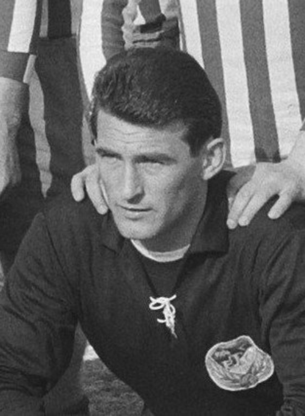 Schwarzes und weißes Foto eines Mannes auf einem grünen Fußballfeld sitzend, der ein T-Shirt und eine Kette trägt, mit zwei stehenden Personen hinter ihm.