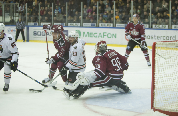 Gruppe von Menschen, die Hockey auf einem Eisring spielen, mit Torpfosten auf der rechten Seite, tragen Helme und halten Stöcke, Zuschauer in Tribünen mit Bannern im Hintergrund.