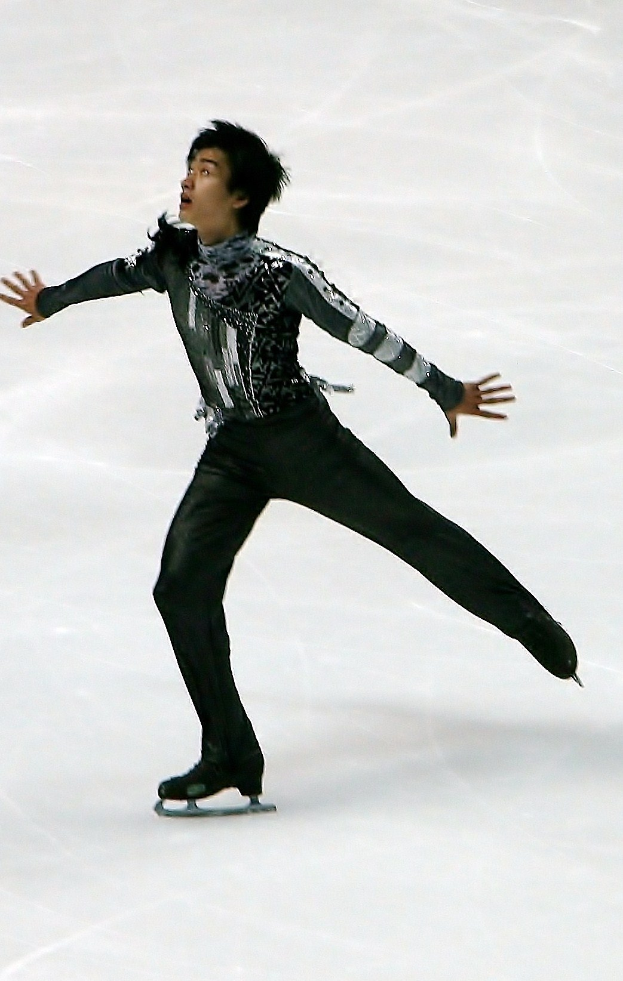 Männlicher Eiskunstläufer in schwarzer Kleidung gleitet über eine Eisfläche und führt eine Choreographie mit synchronen Arm- und Beinbewegungen aus.