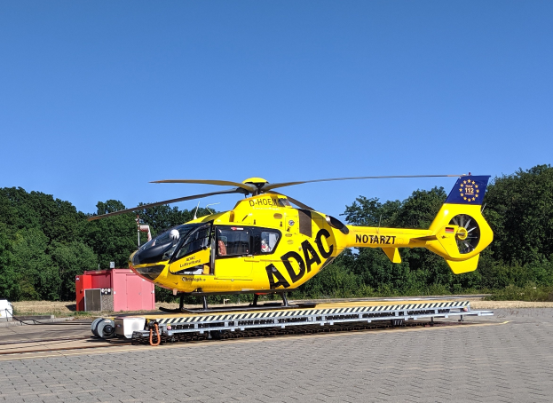 Gelber Hubschrauber auf einem Flughafenvorfeld mit umliegenden Bäumen und wolkenlosem Himmel, wahrscheinlich ein Luftrettungshubschrauber in Deutschland.