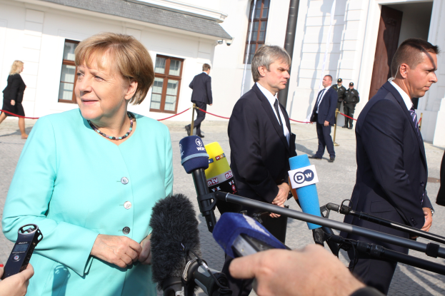 Bundeskanzlerin Angela Merkel spricht zu Reportern vor dem Kanzleramt in Berlin, umgeben von Menschen, einige halten Mikrofone, mit Gebäuden im Hintergrund.