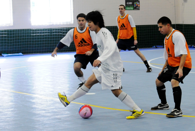 Eine Gruppe junger Männer, die Futsal auf einem Indoor-Court spielt, mit einem Ball in der Mitte und einem Netz im Hintergrund.