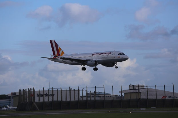Germanwings Airbus A320-200 beim Abheben vom Frankfurter Airport mit Gras darunter, einem Zaun im Vordergrund, Gebäuden und Bäumen im Hintergrund und einer bewölkten Himmel.