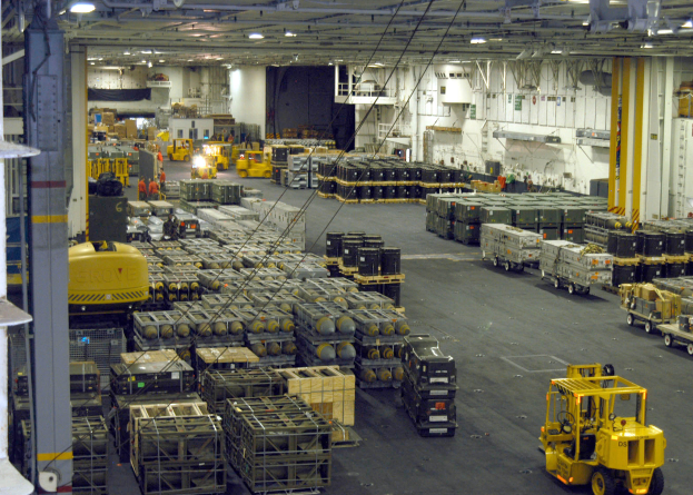 Ein gro├če Lagerhalle mit vielen Lastwagen, Containern, Menschen, Lichtern, Stangen und einem prominenten Rad.