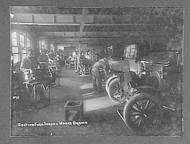 Schwarz-weiß-Foto von Männern, die an einem Auto in einer Ford-Werkstatt arbeiten, mit Fahrzeugen, Werkzeugen und Fenstern im Hintergrund.