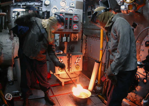 Zwei Männer in Jacken und Helmen arbeiten an einer Maschine in einer Fabrik, einer hält ein Objekt; ein Feuer brennt vor ihnen, und Messgeräte und andere Ausrüstung sind im Hintergrund zu sehen.