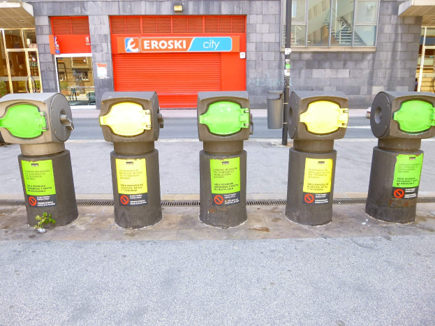 Eine Reihe von Parkautomaten entlang einer Straße mit einem Gebäude mit Glasfenstern und einem Rollladen mit Text sowie einem nahen Müllcontainer.
