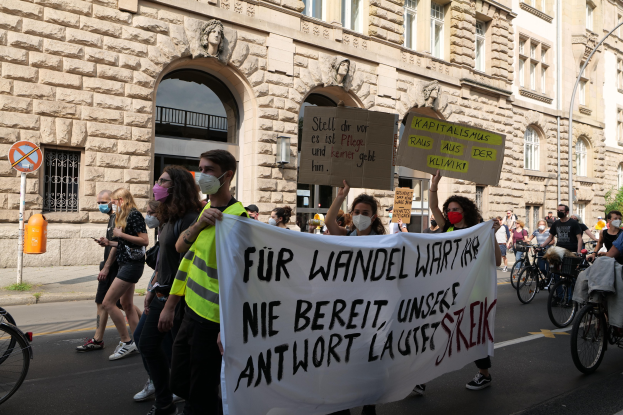 Eine Gruppe von Menschen marschiert durch eine Straße, einige halten Protestschilder und andere fahren Fahrräder, im Hintergrund ein Gebäude mit Fenstern, Bögen, Säulen und Skulpturen.
