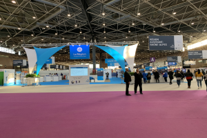 Ein geschäftiges Messestände am Pariser Internationalen Handelsfair 2018, mit Menschen, die herumlaufen, einige Taschen tragend, Banner von der Decke hängend, Tische mit verschiedenen Gegenständen und Überkopfbeleuchtung.