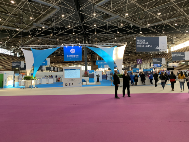 Ein geschäftiges Messestände am Pariser Internationalen Handelsfair 2018, mit Menschen, die herumlaufen, einige Taschen tragend, Banner von der Decke hängend, Tische mit verschiedenen Gegenständen und Überkopfbeleuchtung.