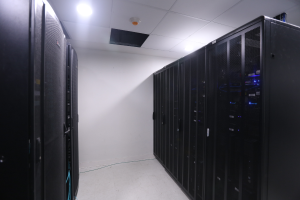 Eine Reihe von Servern in einem Rechenzentrum mit einer Wand im Hintergrund und Leuchten oben, umgeben von diverser Datenspeicherausrüstung.