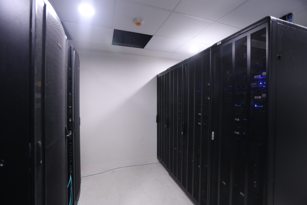 Eine Reihe von Servern in einem Rechenzentrum mit einer Wand im Hintergrund und Leuchten oben, umgeben von diverser Datenspeicherausrüstung.