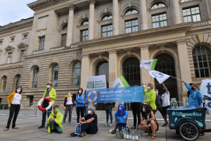 Gruppe von Menschen mit Masken, die Schilder vor einem Gebäude mit Säulen und Bögen während eines Klimastreiks in Berlin halten, mit einem Fahrzeug und einem Geländer im Hintergrund.