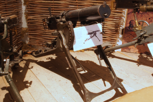 Eine Gatling Gun, eine Art Maschinengewehr, das von der britischen Armee während des Zweiten Weltkriegs verwendet wurde, wird auf einem Holzfußboden in einem Museum ausgestellt, mit einer Wand im Hintergrund und einer Texttafel rechts.