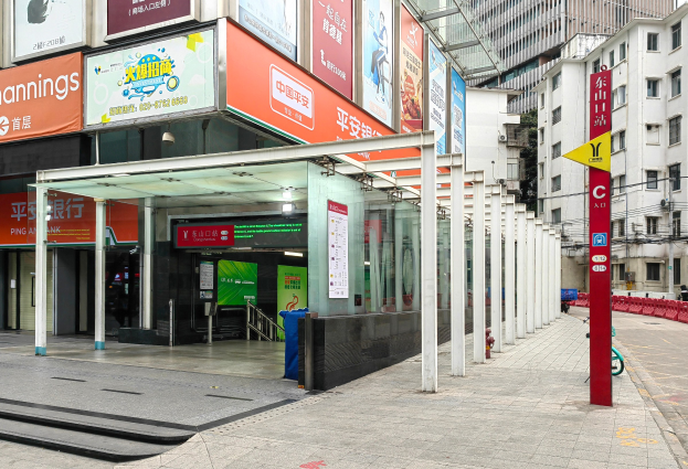 Eine Stadtstraße mit einem Bushaltestelle, Gebäude auf beiden Seiten, eine Straße mit einem Müllcontainer, rote und gelbe Schilder mit Text, Fußgänger auf dem Gehweg und eine Rolltreppe auf der linken Seite.