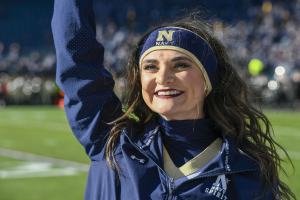 Frau in blauer Jacke und Headband auf einem Footballfeld, die winkend in die Menge lächelt, wahrscheinlich Mitglied der Navy-Frauen-Footballmannschaft.