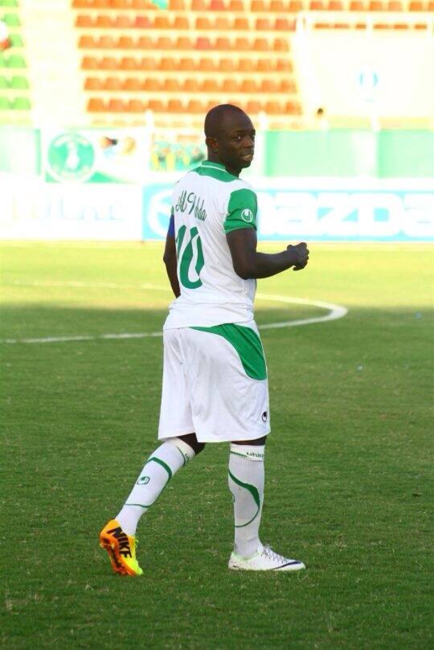 Ein Mann in einem grün-weißen Fußballtrikot feiert ein Tor auf einem Feld, umgeben von Werbetafeln und Stühlen, wahrscheinlich nach dem Sieg in der afrikanischen Nationenmeisterschaft.