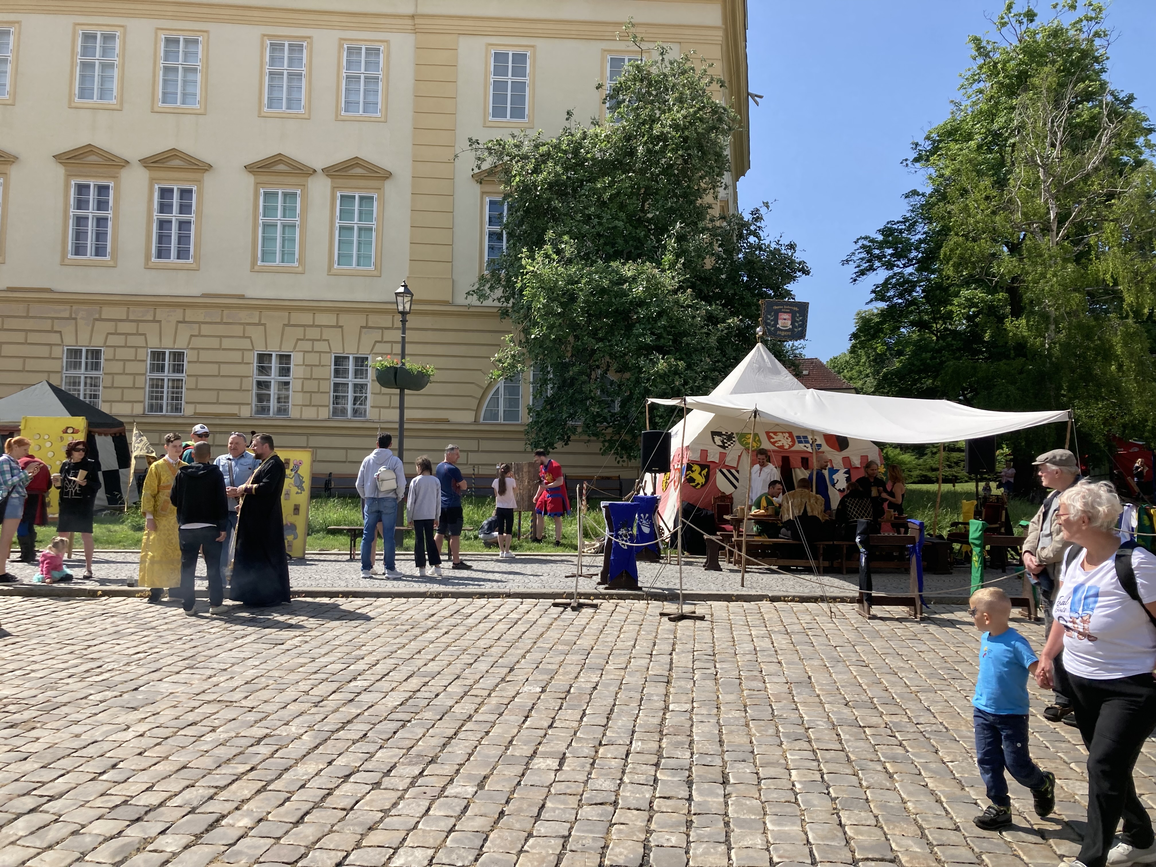 Eine Gruppe von Menschen geht eine Kopfsteinpflasterstraße in Krakau, Polen, entlang, vorbei an einem Gebäude mit Fenstern, Bäumen, Laternen und Zelten, wobei einige eine Mütze und eine Tasche tragen und Tische mit verschiedenen Gegenständen darauf stehen.