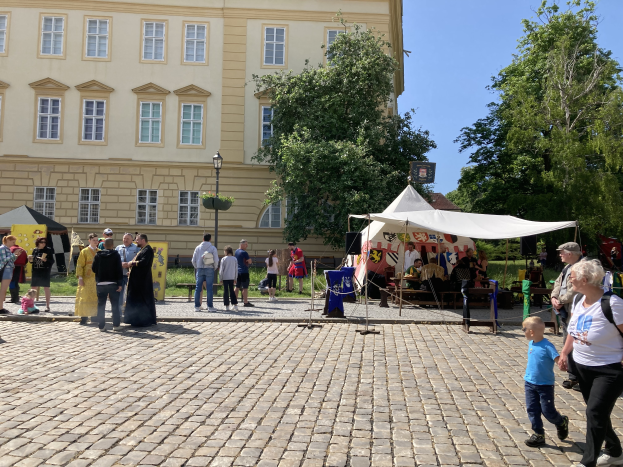 Eine Gruppe von Menschen geht eine Kopfsteinpflasterstraße in Krakau, Polen, entlang, vorbei an einem Gebäude mit Fenstern, Bäumen, Laternen und Zelten, wobei einige eine Mütze und eine Tasche tragen und Tische mit verschiedenen Gegenständen darauf stehen.