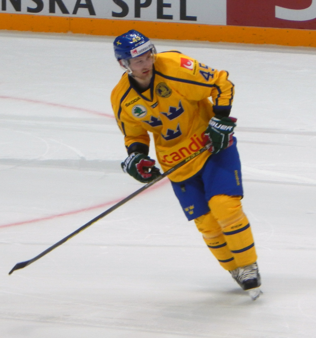 Ein Eishockeyspieler in Bewegung auf dem Eis, der einen Helm, Handschuhe und Schlittschuhe trägt und einen Hockeystock hält, mit einem Hintergrundboard mit Text.