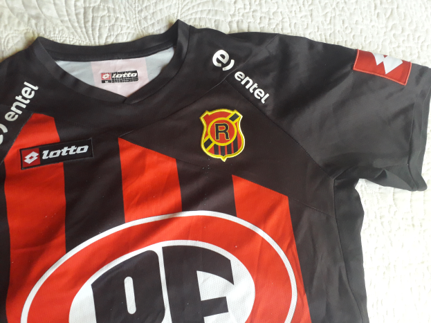 Ein schwarzes und rotes Fußballtrikot mit einem roten und schwarzen Streifen, einem Logo und Text auf einer weißen Oberfläche.