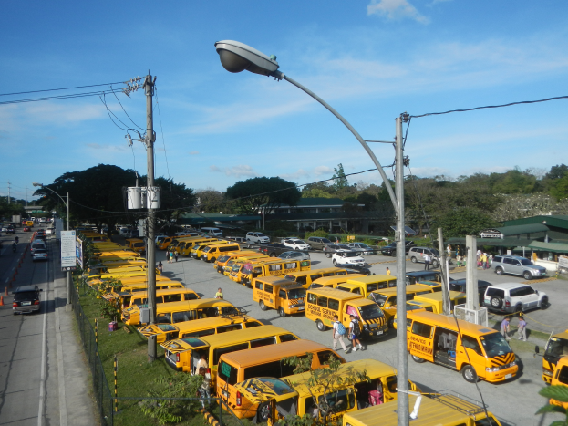 Eine große Anzahl von gelben Schulbussen am Straßenrand geparkt, mit Menschen auf dem Gehweg, Strommasten mit Drähten, Laternenmasten, Bäume, Gebäude und ein bewölkter Himmel im Hintergrund.