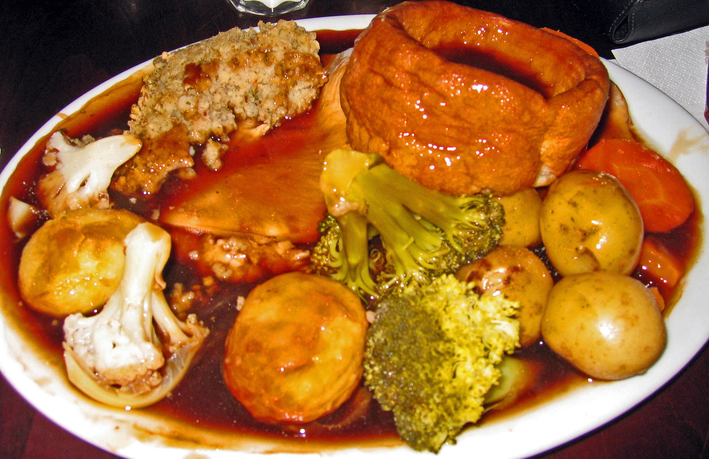 Ein weißer Teller mit Yorkshire-Pudding und Gemüse wie Karotten, Kartoffeln und Zwiebeln, umgeben von Gläsern und Taschentüchern auf einem Tisch.