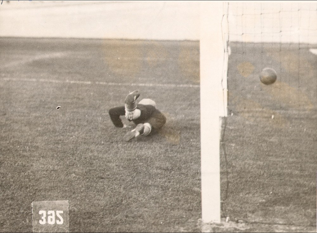 Schwarzes und weißes Foto eines Fußballspielers in der Mitte des Spiels, der einen Ball in der Nähe eines Torpfostens mit Netz auf der rechten Seite fängt.