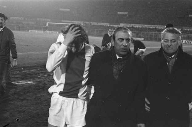 Eine Gruppe von besorgten Männern in Schwarz-Weiß-Kleidung steht zusammen auf einem Fußballfeld, im Hintergrund ist ein Stadion zu sehen, wahrscheinlich nach einem gewonnenen Spiel.