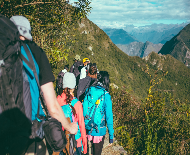 Gruppe von Menschen, die einen Berg hinaufwandern, Rucksäcke und Hüte tragen, mit bewölktem Himmel und einer Landschaft aus Bäumen und Hügeln.